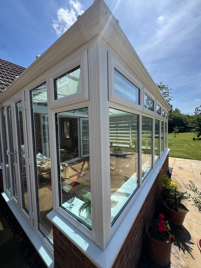 Modern sunroom with large windows and light grey frame attached to brick house, overlooking green garden on sunny day