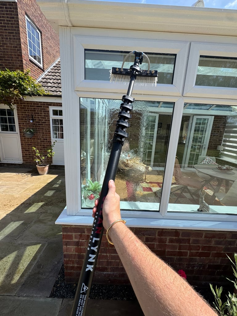 Person holding extendable water-fed pole with brush head for cleaning white uPVC patio doors on brick house
